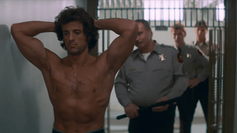 Rambo: First Blood - Sylvester Stallone als Rambo steht mit erhobenen Armen und dem Rücken zu Polizisten zugewandt.