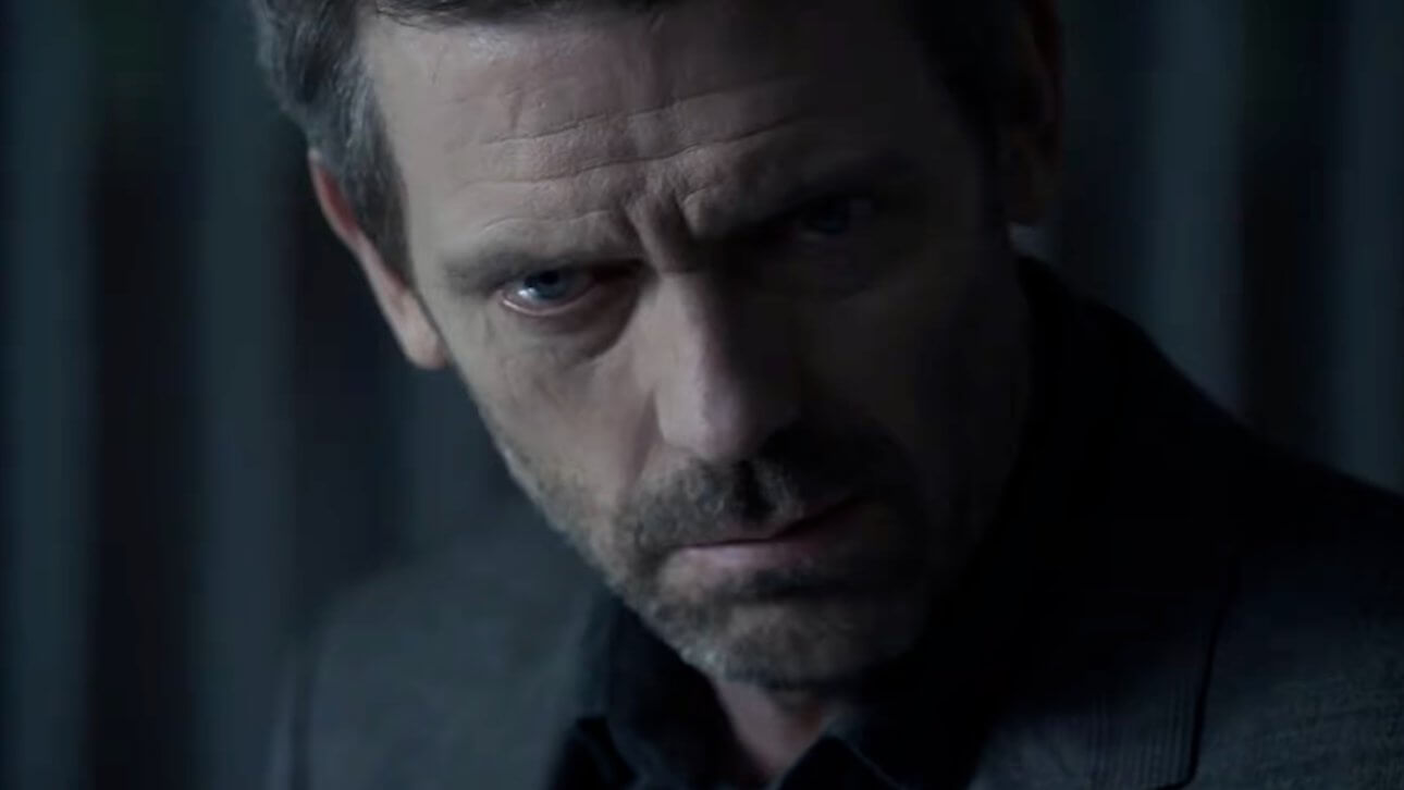 Dr. House: der Schauspieler Hugh Laurie sitzt im Halbdunklen. Die Hälfte seines Gesichts ist von einem Schatten bedeckt. Er wirkt besorgt.