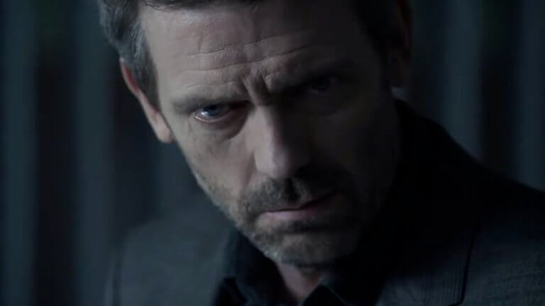 Dr. House: der Schauspieler Hugh Laurie sitzt im Halbdunklen. Die Hälfte seines Gesichts ist von einem Schatten bedeckt. Er wirkt besorgt.