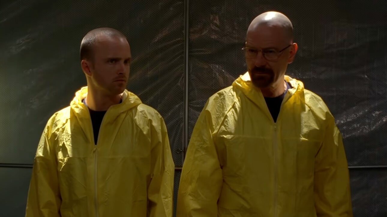 Breaking Bad: Walter White und sein Compagnon tragen gelbe Schutzanzüge und stehen in einem schlecht beleuchteten Lagerraum.
