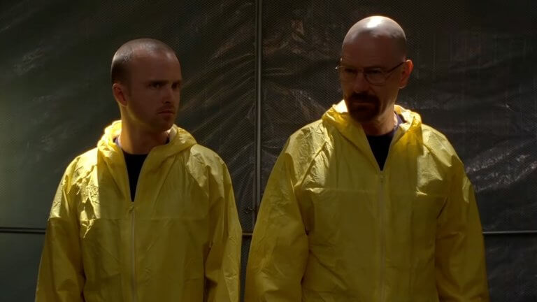 Breaking Bad: Walter White und sein Compagnon tragen gelbe Schutzanzüge und stehen in einem schlecht beleuchteten Lagerraum.