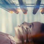 DISCLOSURE DAY: Der Tag der Wahrheit: ein Mädchen scheint von Aliens untersucht zu werden. Der Raum ist sehr hell. Über ihrem Kopf schwebt eine seltsame Apparatur, und eine Hand mit drei langen Fingern hält den Kopf fest.