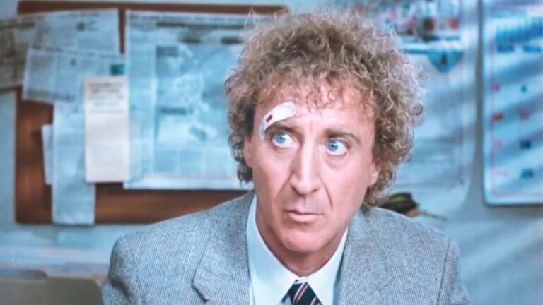 Die Frau in Rot: Gene Wilder sitzt im Büro und macht sich Gedanken. Er trägt einen grauen Anzug und hat ein Pflaster über dem rechten Auge.