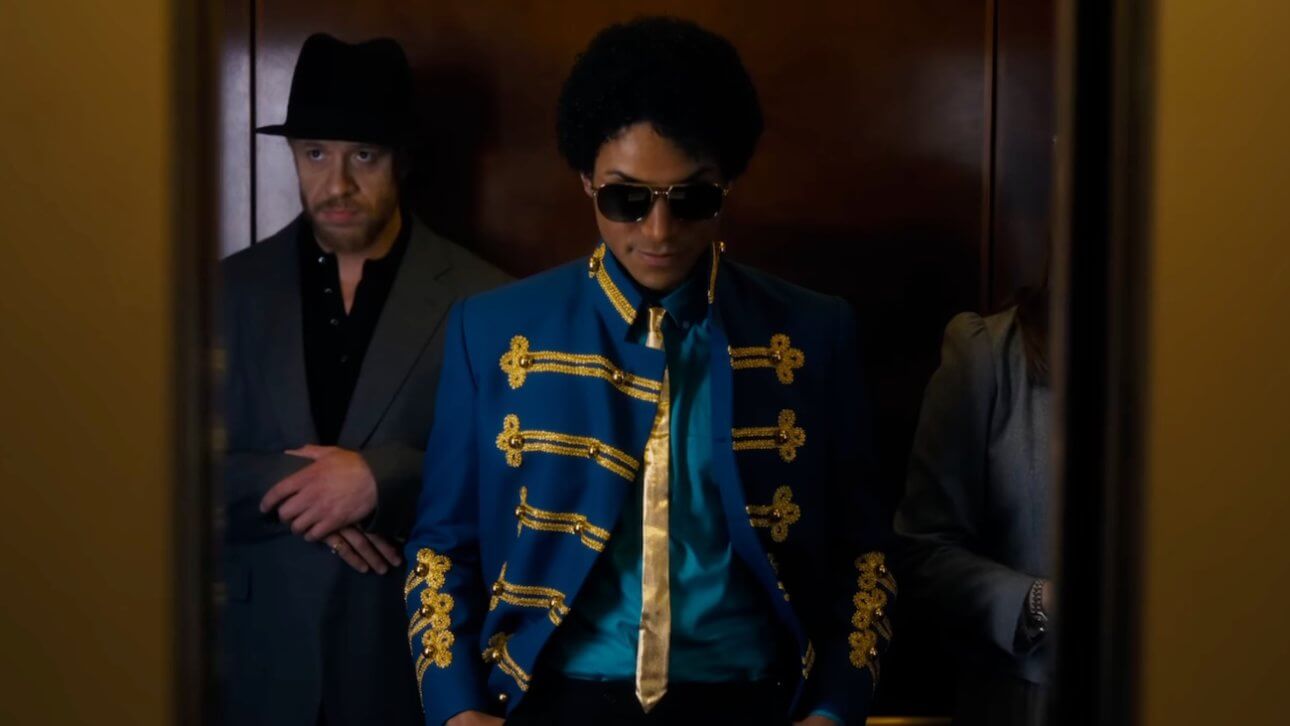 Michael: Michael Jackson steht mit seiner Crew im Aufzug. Er trägt ein auffälliges Oberteil, das an einen Zirkusdirektor erinnert.