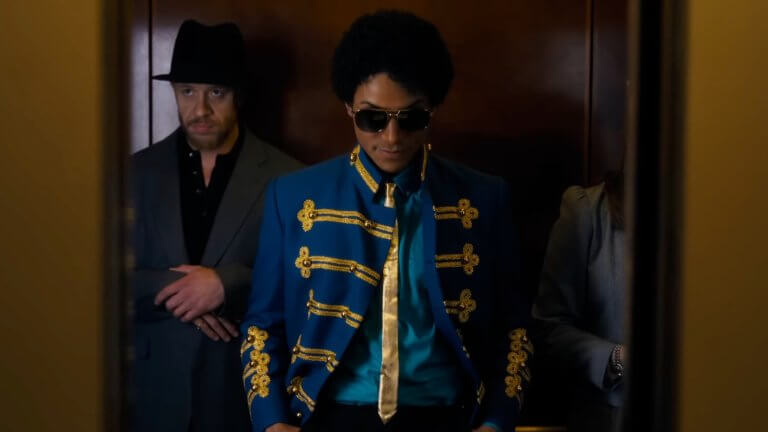 Michael: Michael Jackson steht mit seiner Crew im Aufzug. Er trägt ein auffälliges Oberteil, das an einen Zirkusdirektor erinnert.