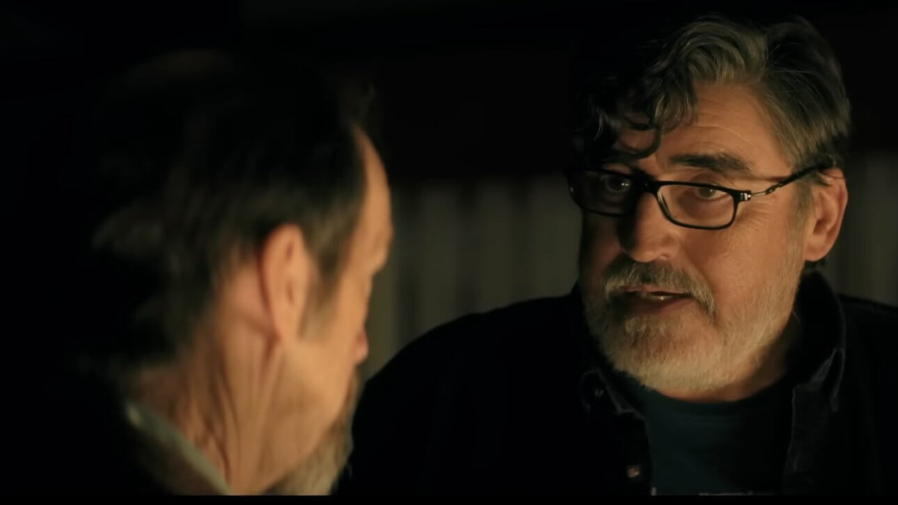 The Boroughs: Alfred Molina trägt eine Brille und steht im Halbdunkeln. Ihm gegenüber befindet sich eine andere Person mit dem Rücken zur Kamera.