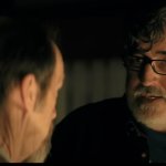 The Boroughs: Alfred Molina trägt eine Brille und steht im Halbdunkeln. Ihm gegenüber befindet sich eine andere Person mit dem Rücken zur Kamera.
