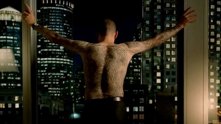 Prison Break: ein tätowierter Mann steht vor dem Fenster mit ausgestreckten Armen. Die Kamera zeigt seinen tätowierten Rücken. Im Hintergrund ist die Skyline einer Stadt zu erkennen.