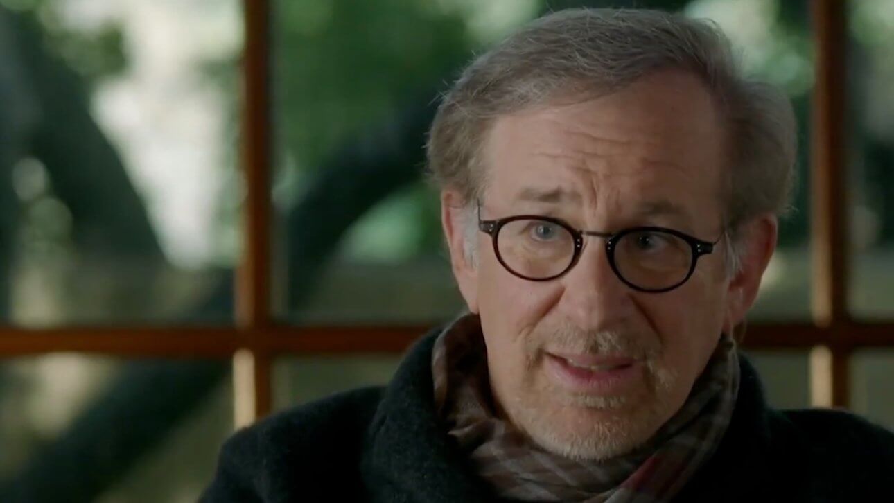 Spielberg (2017) HBO Documentary: Steven Spielberg sitzt vor einem Fenster. Im Hintergrund ist verschwommen ein Garten zu erkennen. Er trägt eine Brille und einen Schal.