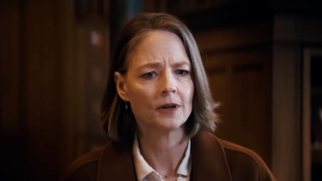 In einer Szene aus „A PRIVATE LIFE“ steht Jodie Foster in einer Bibliothek und trägt einen braunen Mantel. Die Aufnahme zeigt sie von den Schultern aufwärts.