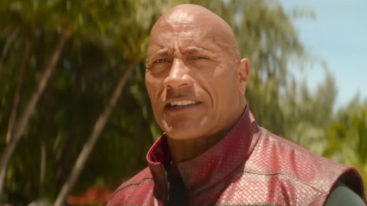 RED ONE: Dwayne "The Rock" Johnson trägt eine schwarz-rote Weste und steht am Strand. Im Hintergrund sind Palmen zu sehen.