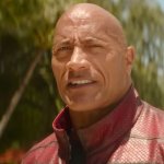 RED ONE: Dwayne "The Rock" Johnson trägt eine schwarz-rote Weste und steht am Strand. Im Hintergrund sind Palmen zu sehen.