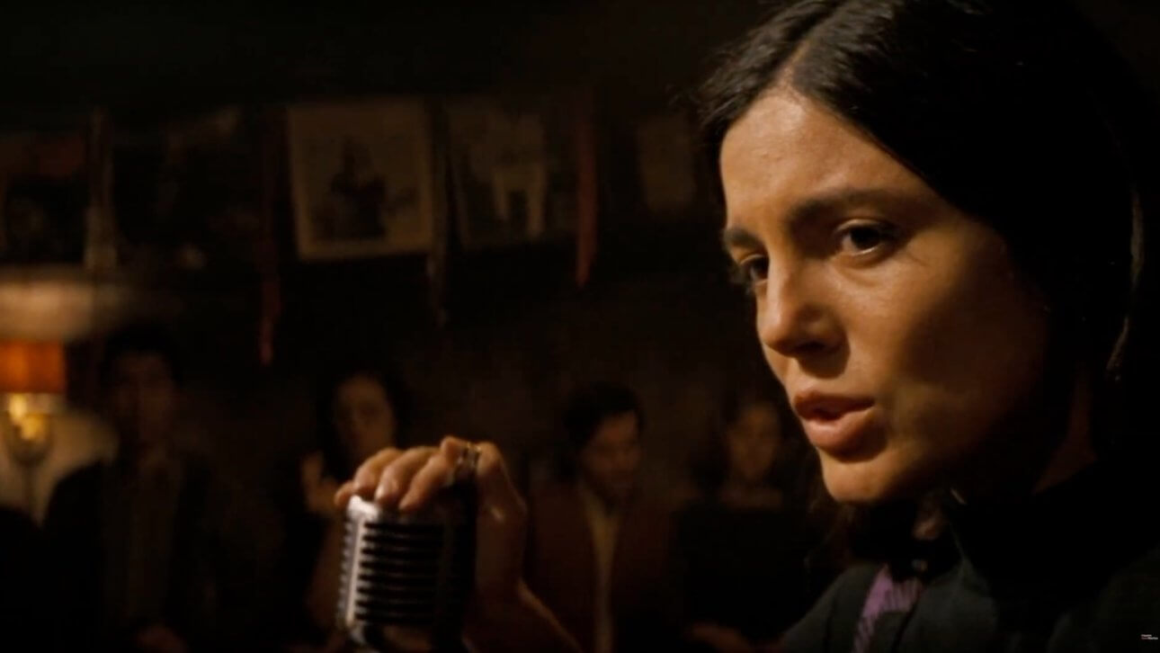 A Complete Unknown: Monica Barbaro als Joan Baez sitzt auf der Bühne und singt einen Song. Wir sehen ihren Kopf und ihre Hand, die das Mikrofon berührt.