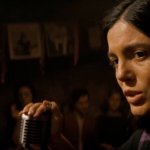 A Complete Unknown: Monica Barbaro als Joan Baez sitzt auf der Bühne und singt einen Song. Wir sehen ihren Kopf und ihre Hand, die das Mikrofon berührt.