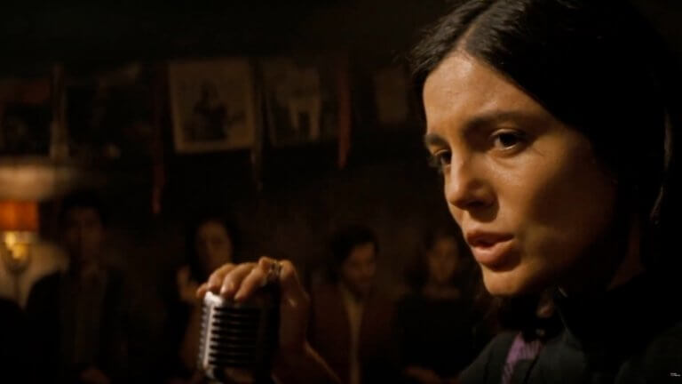 A Complete Unknown: Monica Barbaro als Joan Baez sitzt auf der Bühne und singt einen Song. Wir sehen ihren Kopf und ihre Hand, die das Mikrofon berührt.