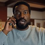 Yahya Abdul-Mateen II telefoniert in der Rolle des Simon Williams und blickt überrascht. Im Hintergrund ist ein Raum mit mehreren Gemälden zu erkennen.