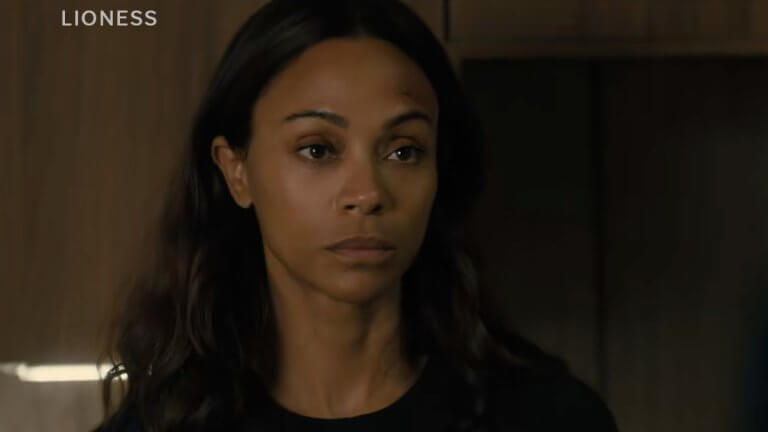 Lioness: Die Schauspielerin Zoe Saldana steht vor einem dunkelbraunen Schrank. Im Vordergrund ist der schemenhafte Umriss einer blonden Frau erkennbar.