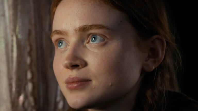 Stranger Things: Sadie Sink blickt durch das Fenster. Neben ihr sind beige Gardinen zu sehen.