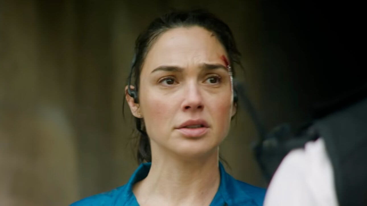 The Runner: Gal Gadot wirkt sichtlich mitgenommen. Sie trägt ein verschmutztes Oberteil in blauer Farbe. Ihr gegenüber erkennt man die Umrisse von zwei britischen Polizisten.