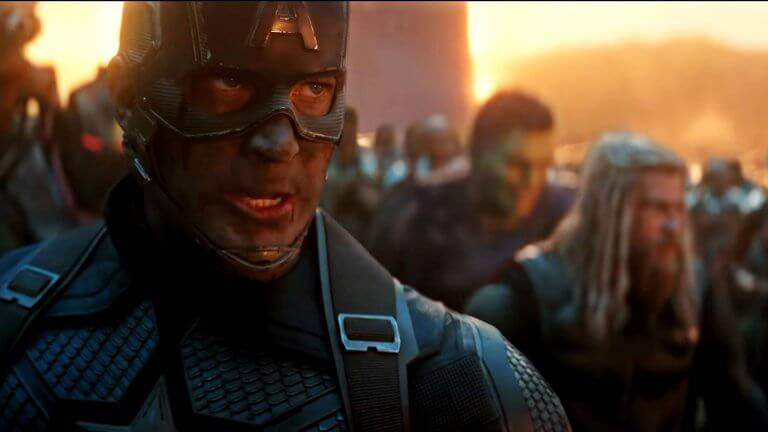 Captain Amercia ist gezeichnet vom Kampf, aber blickt grimmig und entschlossen. Hinter ihm sind eine Menge Charaktere versammelt, unter anderem Hulk und Thor.