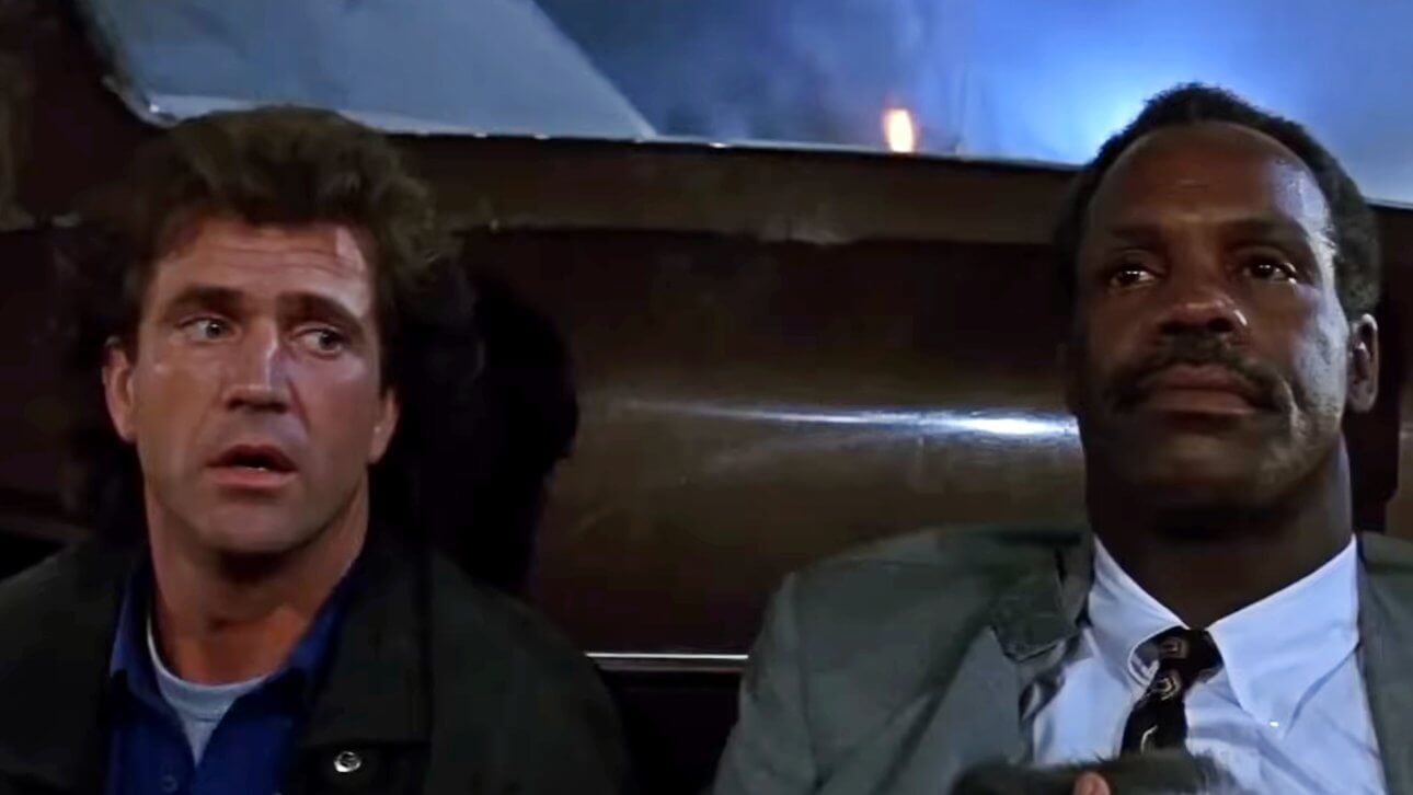 Mel Gibson und Danny Glover verstecken sich hinter einem Auto. Während Gibson schuldbewusst zur Seite blickt, starrt Glover resigniert ins Leere.