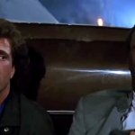 Mel Gibson und Danny Glover verstecken sich hinter einem Auto. Während Gibson schuldbewusst zur Seite blickt, starrt Glover resigniert ins Leere.