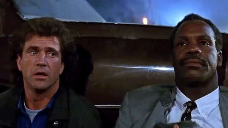 Mel Gibson und Danny Glover verstecken sich hinter einem Auto. Während Gibson schuldbewusst zur Seite blickt, starrt Glover resigniert ins Leere.