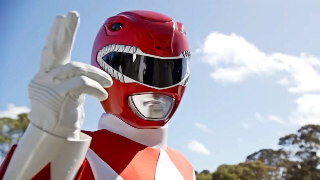 Der rote Power Ranger ist in voller Montur zu sehen. Hinter ihm sind der Himmel und einige Bäume zu sehen, während er die rechte Hand hochhält.