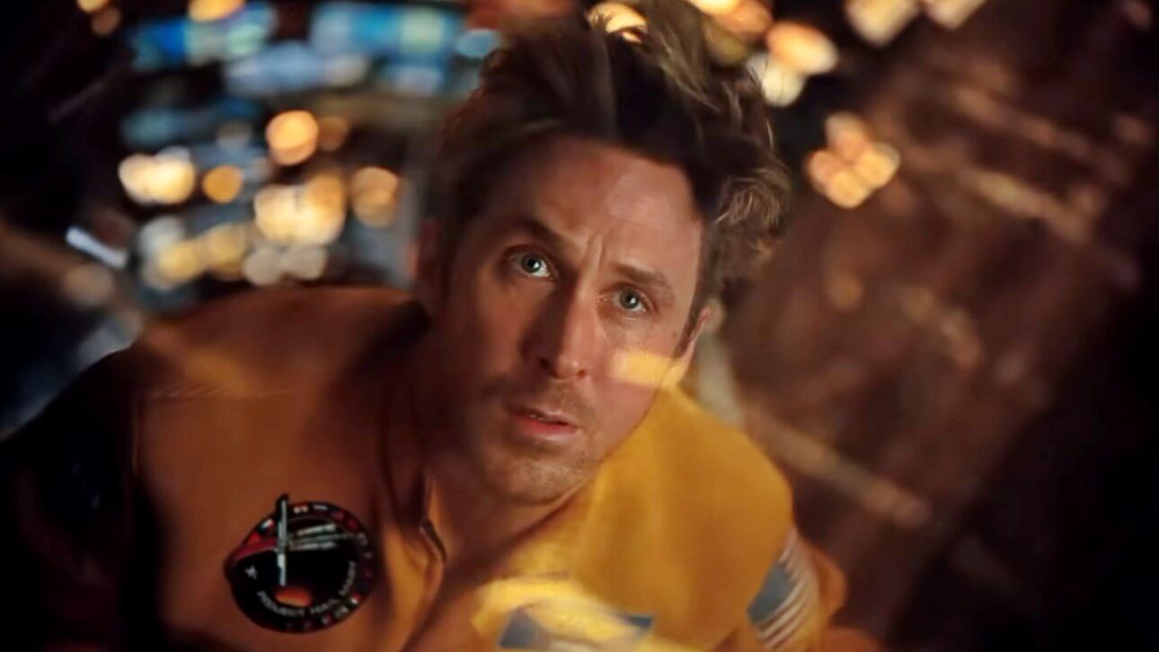 Ryan Gosling schaut nervös zur Kamera. Eine Fensterscheibe vor seinem Gesicht sorgt für die Spiegelung vieler kleiner Lichter. Er trägt eine orangfarbene Astronautenuniform.