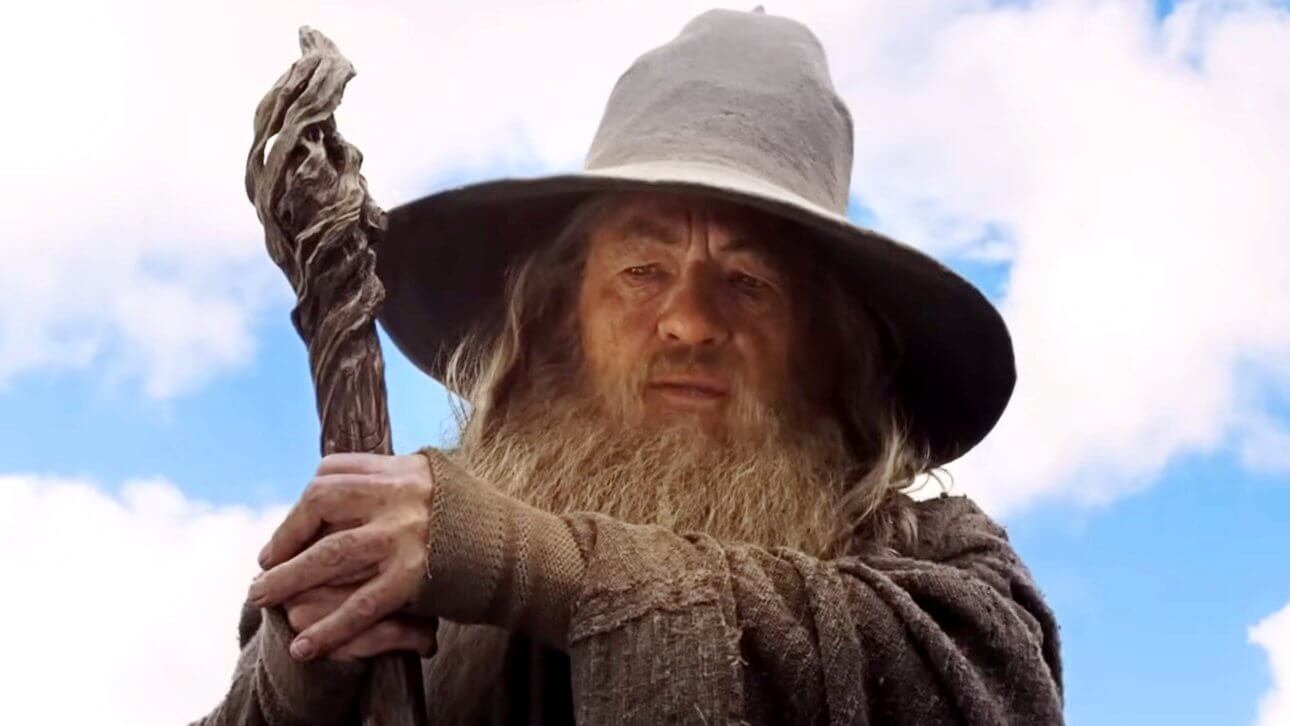 Der Zauberer Gandalf steht vor einem strahlend blauen Himmel. Er trägt das ikonische graue Gewand und hält seinen Holzstab in beiden Händen.