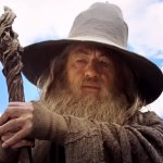 Der Zauberer Gandalf steht vor einem strahlend blauen Himmel. Er trägt das ikonische graue Gewand und hält seinen Holzstab in beiden Händen.