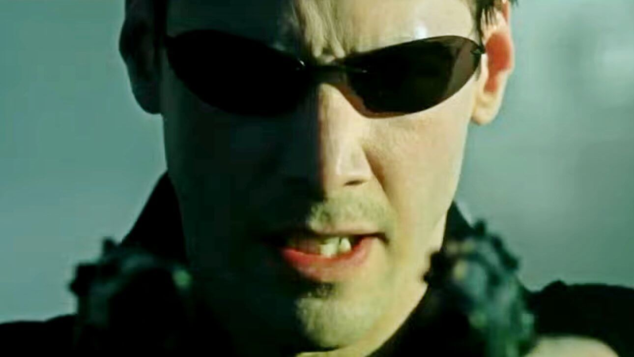 Neo zielt mit zwei Pistolen. Er trägt einen dunklen Mantel und die ikonische Sonnenbrille. Sein Gesichtsausdruck ist angespannt und grimmig.