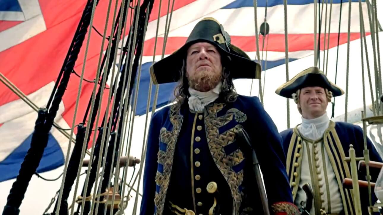 Geoffrey Rush steht als Captain Hector Barbossa in einer prunkvollen Uniform an Bord eines Schiffes. Hinter ihm ist die britische Flagge zu sehen.