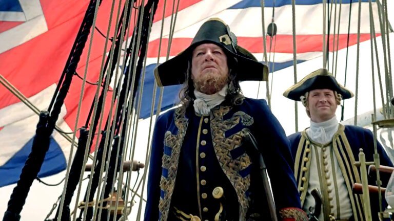 Geoffrey Rush steht als Captain Hector Barbossa in einer prunkvollen Uniform an Bord eines Schiffes. Hinter ihm ist die britische Flagge zu sehen.