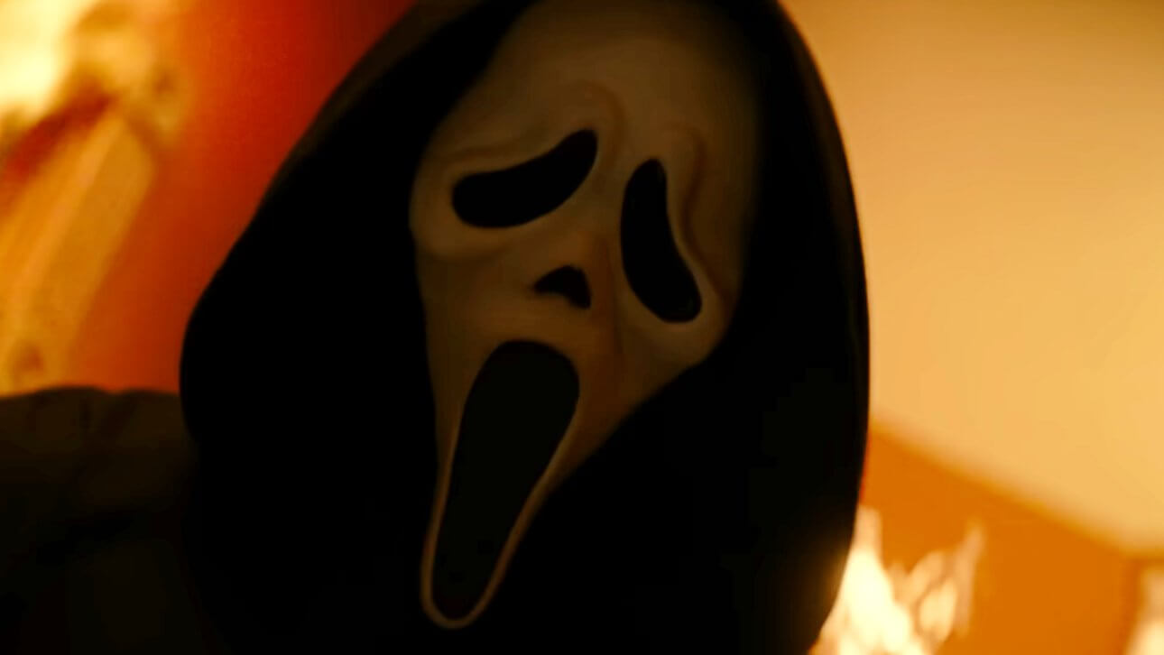Der berühmte Ghostface-Killer steht in einem brennenden Haus. Er trägt die ikonische Maske und den bekannten schwarzen Umhang.