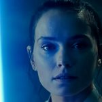 Rey blickt direkt in die Kamera mit einer unergründlichen Miene. Sie hält ein blaues Lichtschwert, welches in der dunklen Umgebung ihre rechte Gesichtshälfte in kaltes Licht taucht.
