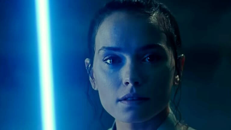 Rey blickt direkt in die Kamera mit einer unergründlichen Miene. Sie hält ein blaues Lichtschwert, welches in der dunklen Umgebung ihre rechte Gesichtshälfte in kaltes Licht taucht.