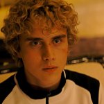 Haymitch Abernathy sitzt in einem schwarz-weißen Trainingsanzug auf einer Couch und blickt angespannt in Richtung der Kamera. Sein gesicht ist von Schrammen und Wunden gezeichnet.