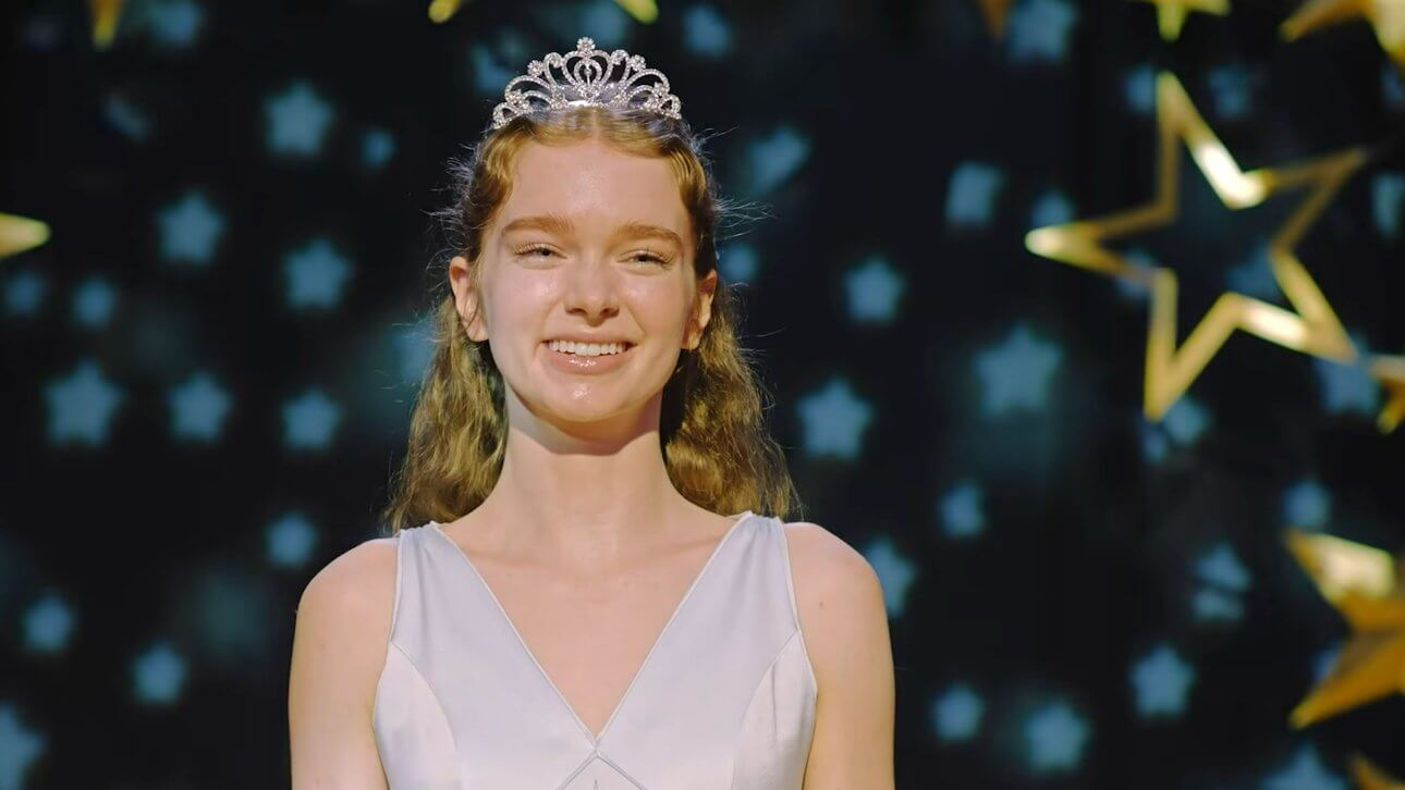 Carrie White steht lächelnd auf einer Bühne. Sie trägt ein weißes Kleid und eine Krone. Hinter ihr ist ein Vorhang mit blauen und goldenen Sternen zu sehen.