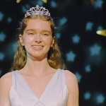 Carrie White steht lächelnd auf einer Bühne. Sie trägt ein weißes Kleid und eine Krone. Hinter ihr ist ein Vorhang mit blauen und goldenen Sternen zu sehen.