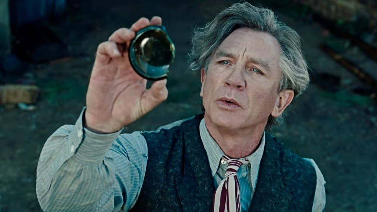 Der Magier, gespielt von Daniel Craig, hält den zerbochenen Boden einer Flasche hoch und blickt fasziniert hindurch. Er trägt Hemd, Krawatte und Weste.