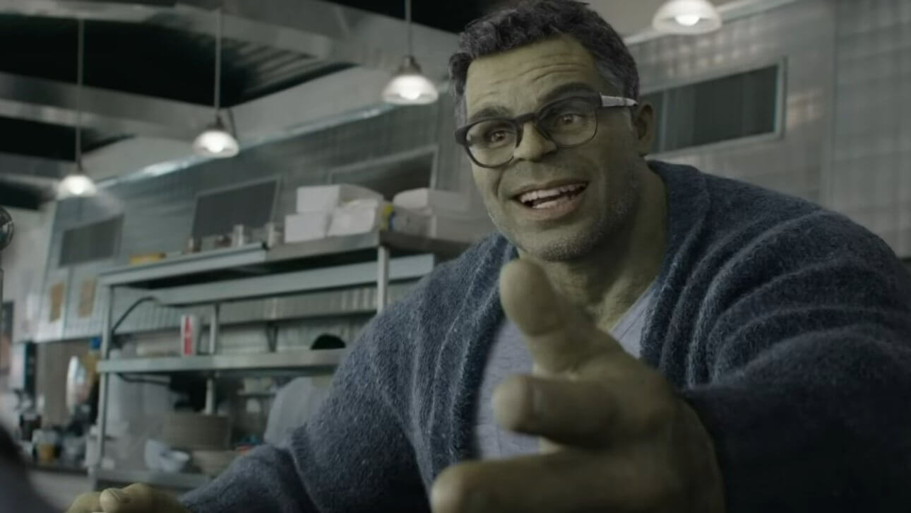 Bruce Banner ist als Professor Hulk zu sehen. Er sitzt in einem Restaurant und gestikuliert beim Reden. Er trägt ein graues T-Shirt, eine Brille und eine dunkelblaue Strickjacke.