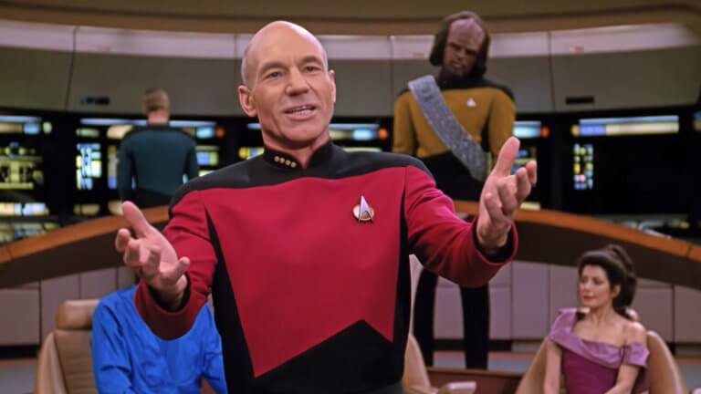 Das Bild zeigt Captain Jean-Luc Picard in einer Uniform auf der Brücke der Enterprise, während er gestikulieren spricht. Im Hintergrund sind weitere Crewmitglieder wie Worf und Counselor Troi zu sehen.