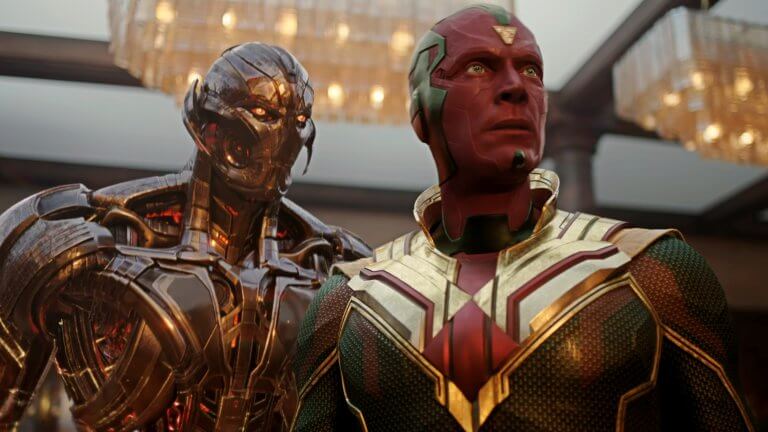 Vision steht mit seinen ikonischen roten, grünen und goldenen Farben in einem Haus. Hinter ihm ist die böse KI Ultron zu sehen, die ihn bedrohlich anstarrt.